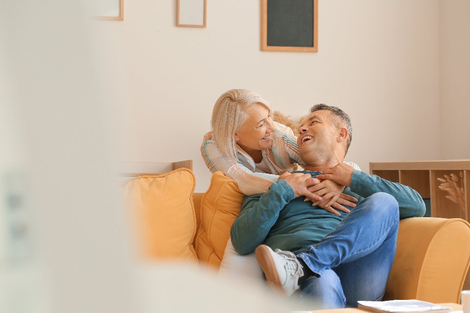 assurance-emprunteur-senior-60-70-ans Couple de seniors souriant, enlacé sur un canapé dans leur salon, illustrant l'assurance emprunteur pour les plus de 60 ans.