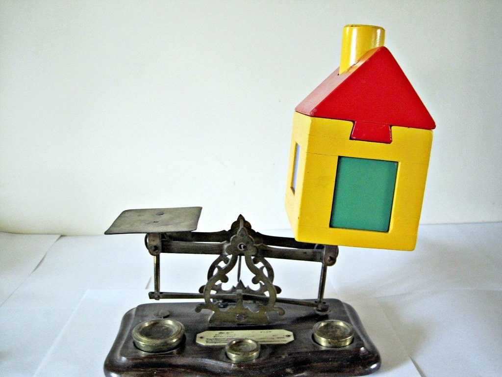comparatif-assurance-emprunteur-grandes-banques Maisonnette miniature en équilibre sur une ancienne balance à poids, symbolisant la comparaison des assurances emprunteur entre plusieurs banques.
