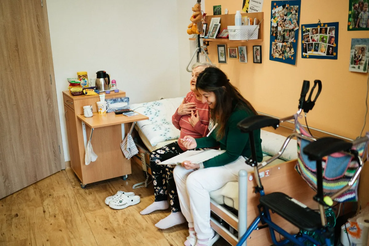 Une femme âgée assise sur un lit d'hôpital dans une chambre individuelle, en conversation avec une infirmière