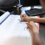 Une femme remplit un formulaire papier à une table, un calendrier mural accroché derrière elle