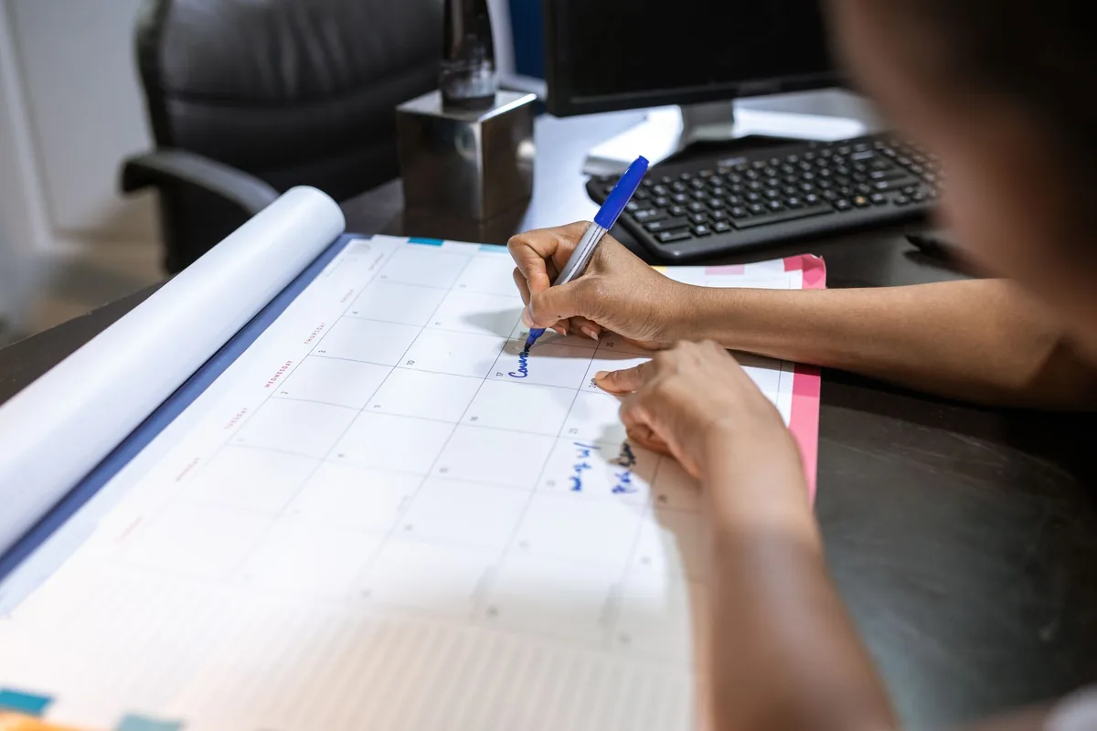Une femme remplit un formulaire papier à une table, un calendrier mural accroché derrière elle